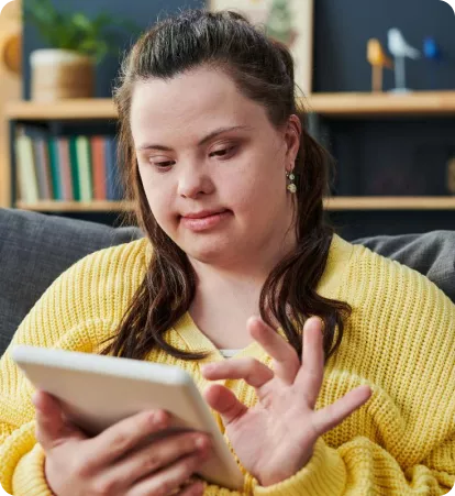 A young woman in a yellow sweater is sitting on a couch, using a tablet and smiling. Bookshelves are visible in the background