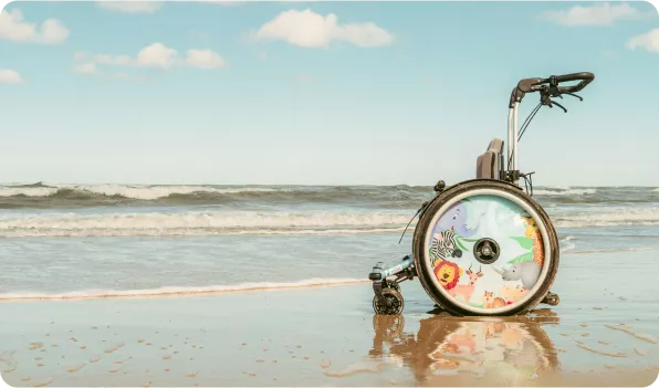 A colorful beach wheelchair is positioned on wet sand, with ocean waves and a clear sky in the background