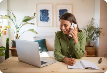A woman in a green shirt is sitting at a desk, talking on the phone while writing in a notebook, with a laptop in front of her