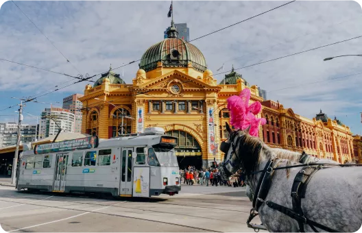 A horse with a pink decoration stands near a tram in front of a grand yellow building, bustling with people