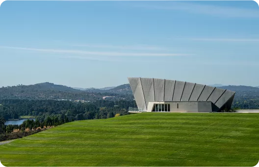 A modern building with a unique angular design sits on a green hillside, overlooking a lake and distant mountains under a clear blue sky