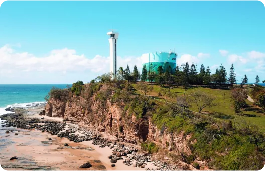 A coastal view featuring a rocky cliff, a tall observation tower, and a green water tank, with the ocean and blue sky in the background
