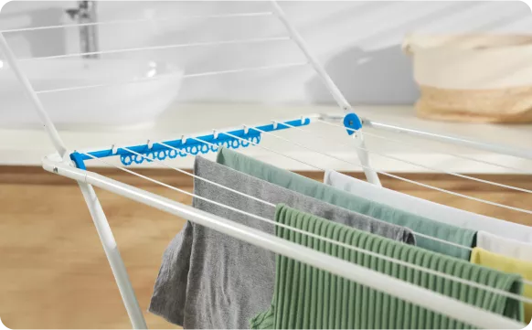 A clothes drying rack with various garments hanging, set in a bright indoor space. A sink and woven basket are visible in the background