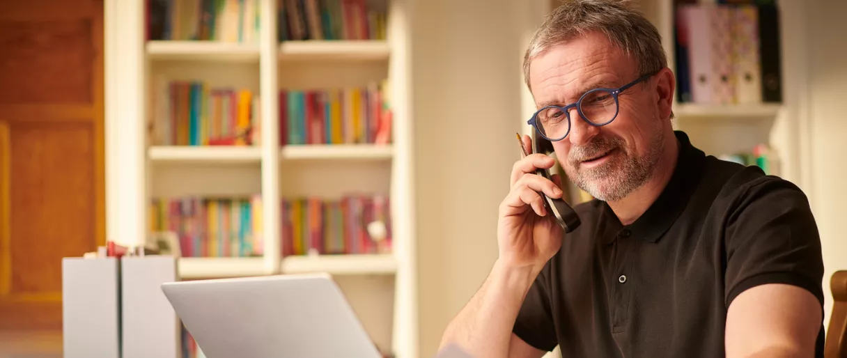A man with glasses is talking on the phone while sitting at a desk with a laptop, surrounded by bookshelves