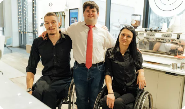 Three individuals pose together in a café setting. Two are in wheelchairs, and all are smiling, dressed in black and white attire