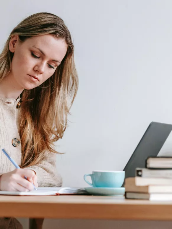 A woman with long hair writes in a notebook at a desk, with a laptop and a cup of tea nearby