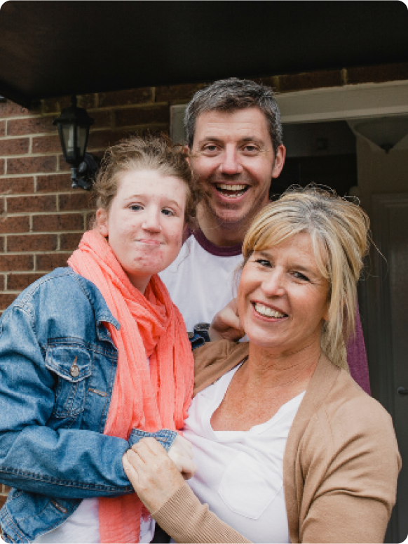 A smiling family poses together outdoors, with a woman and two young people, one wearing a denim jacket and an orange scarf