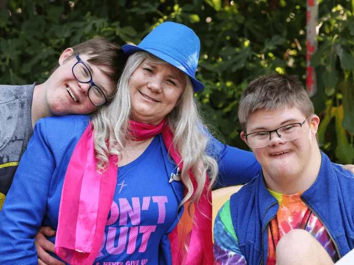 A woman with long hair and a blue hat poses with two smiling young adults, all wearing colorful outfits, against a green background