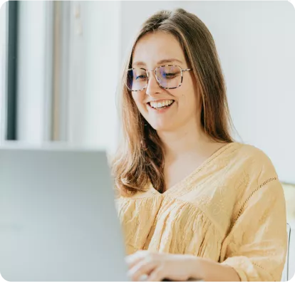 A woman with long hair and glasses smiles while working on a laptop in a bright, modern setting