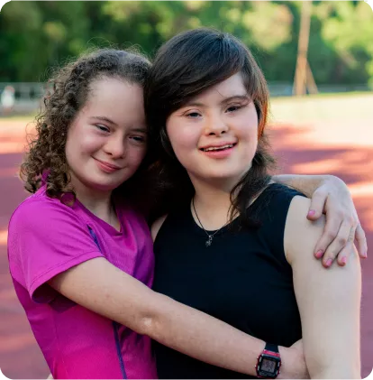 Two girls embrace happily outdoors, smiling at the camera. One wears a pink shirt, and the other a black tank top, with a track background