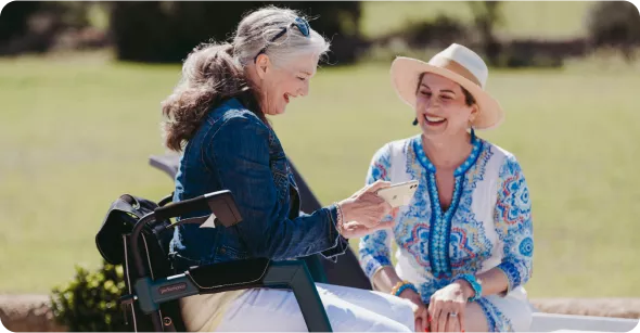 Two women are sitting outdoors, one in a wheelchair, sharing a joyful moment while looking at a phone. Bright, sunny setting