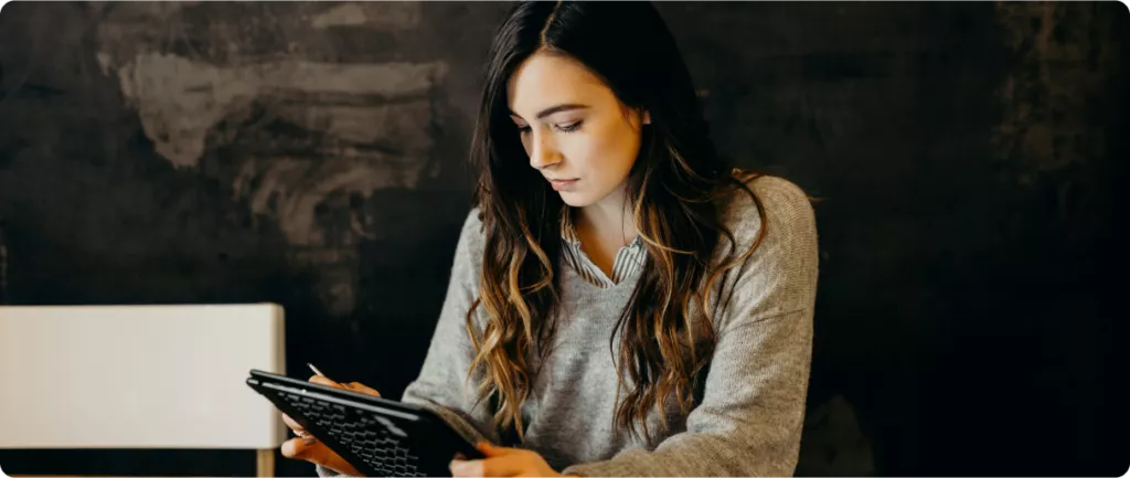 A young woman with long hair sits at a table, focused on a tablet in her hands, against a dark background