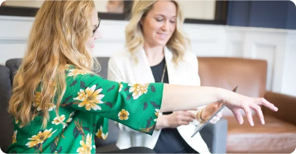 Two women are seated in a cozy setting, engaged in conversation. One is pointing at a tablet while the other smiles, listening attentively