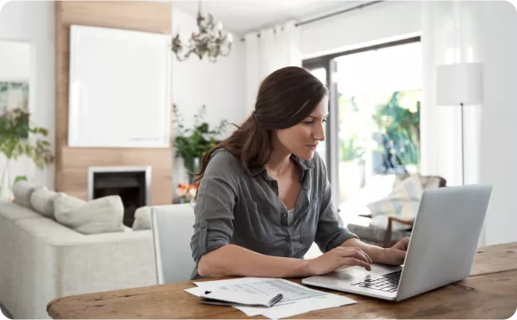 A woman works on a laptop at a wooden table, with papers and a pen nearby, in a bright, modern living room