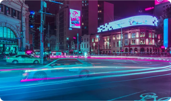 A vibrant city scene at night, featuring moving cars with light trails, illuminated buildings, and colorful billboards