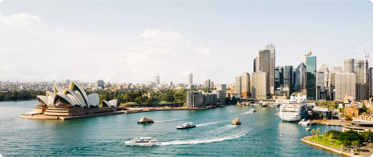 A scenic view of Sydney's waterfront featuring the iconic Opera House and a skyline of modern buildings. Boats navigate the water