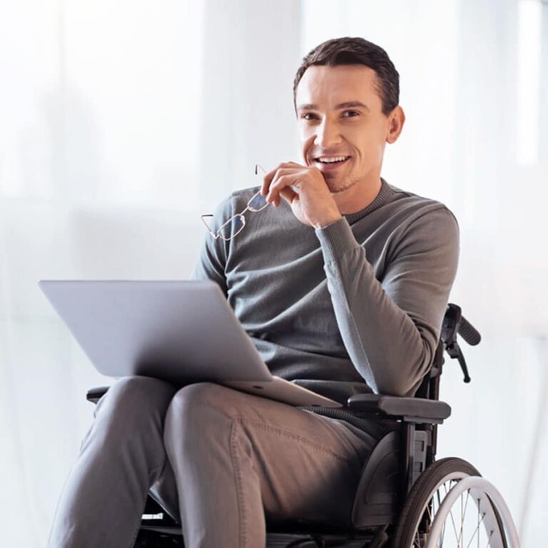 A man in a wheelchair is smiling while holding glasses and using a laptop. He wears a gray sweater and sits in a bright, modern space