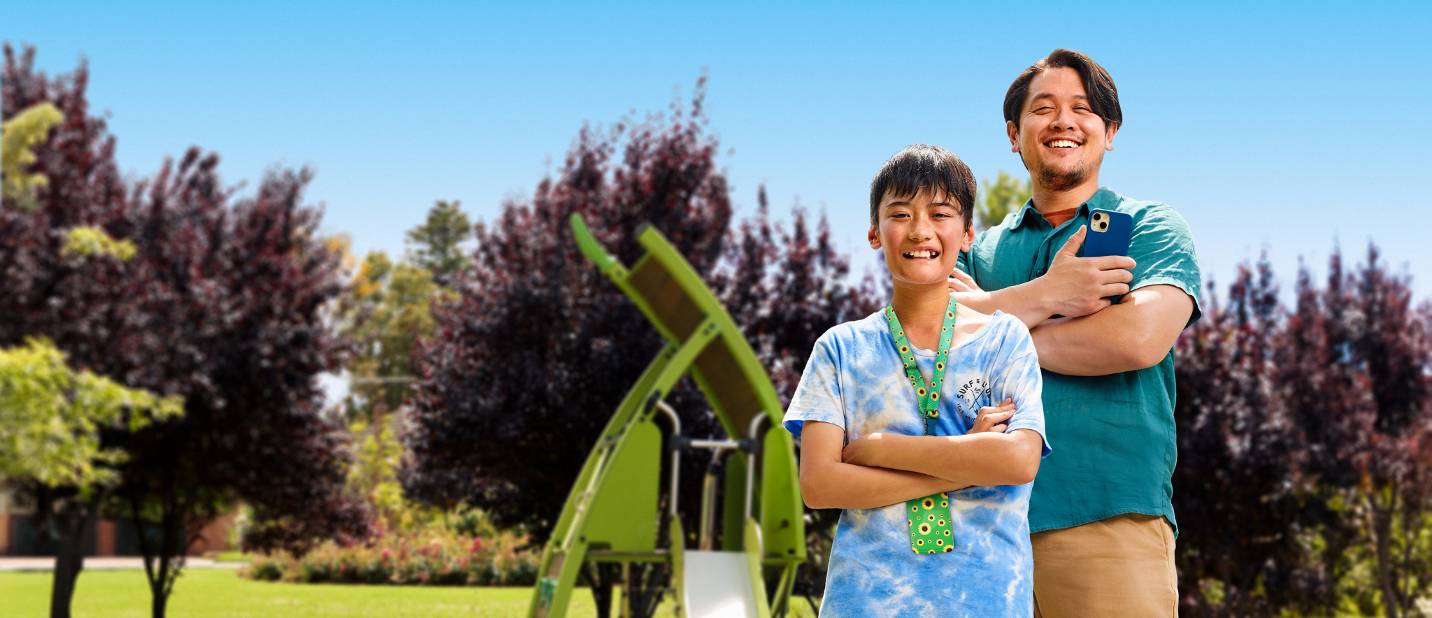 Two smiling individuals stand in a park with green trees and a playground in the background. One is a child wearing a colorful shirt, and the other is an adult holding a phone