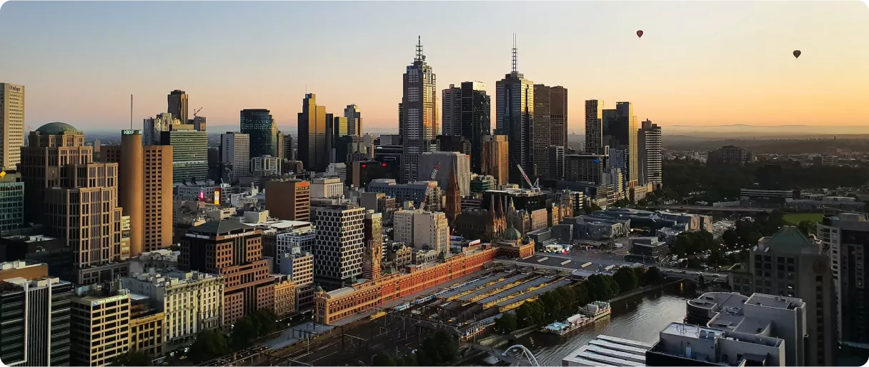 A panoramic view of a city skyline at sunset, featuring tall buildings and hot air balloons in the sky