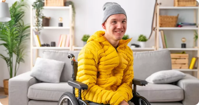 A smiling person in a yellow puffer jacket and gray beanie sits in a wheelchair, in a cozy indoor setting