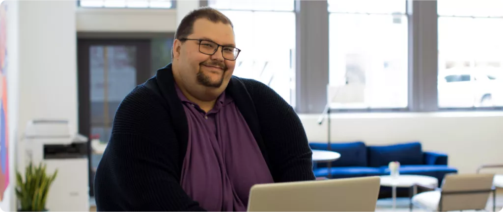 A person with glasses sits at a table with a laptop, smiling in a bright, modern office space