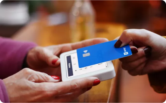 A person is using a blue card to make a payment on a small card reader, with another hand holding the reader