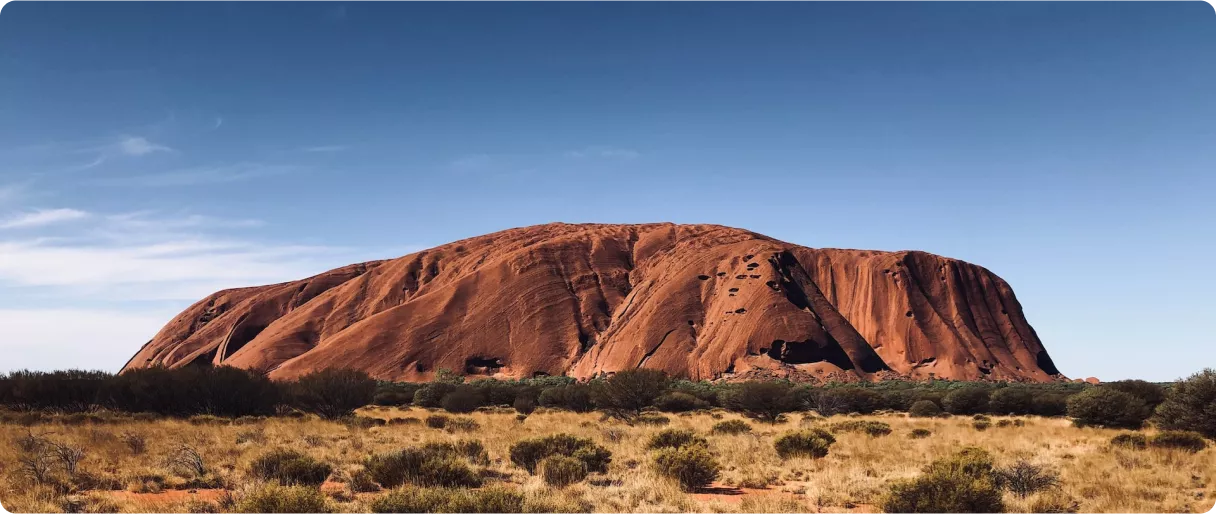 A large, reddish rock formation rises against a clear blue sky, surrounded by sparse vegetation and open land