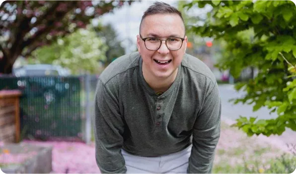 A smiling person in a green long-sleeve shirt leans forward outdoors, surrounded by blooming trees and scattered pink petals