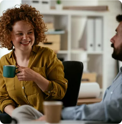 A woman with curly hair smiles while holding a mug, engaging in conversation with a man in a casual office setting