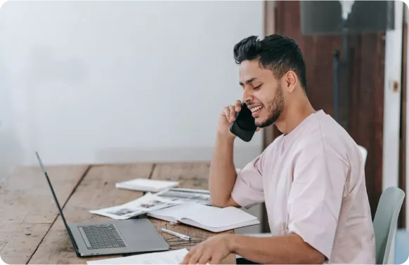 A man in a light pink shirt is smiling while talking on the phone at a wooden desk with a laptop and papers