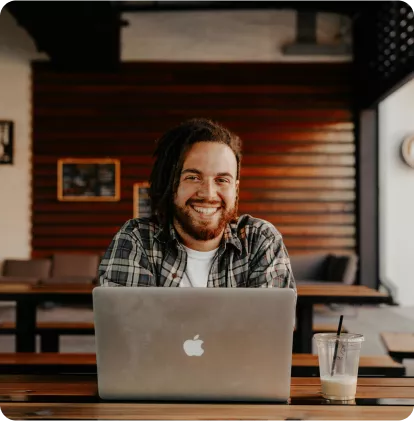 A smiling person with dreadlocks sits at a table with a laptop and a drink, in a cozy indoor setting