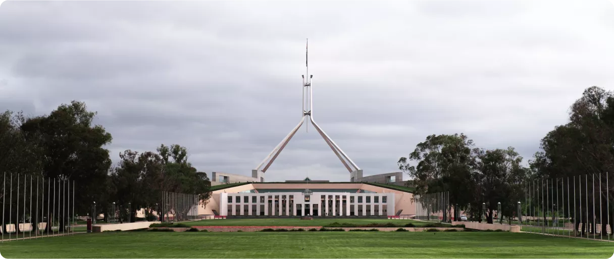 A wide view of a modern building with a tall spire, surrounded by green grass and trees under a cloudy sky
