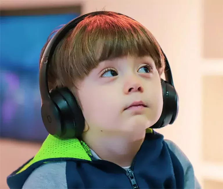 A young child wearing black headphones looks up thoughtfully, dressed in a blue and green outfit