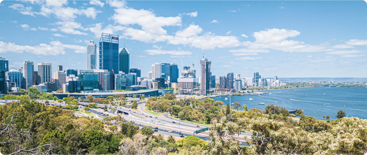 A panoramic view of a city skyline with modern skyscrapers, lush greenery, and a river under a bright blue sky with scattered clouds