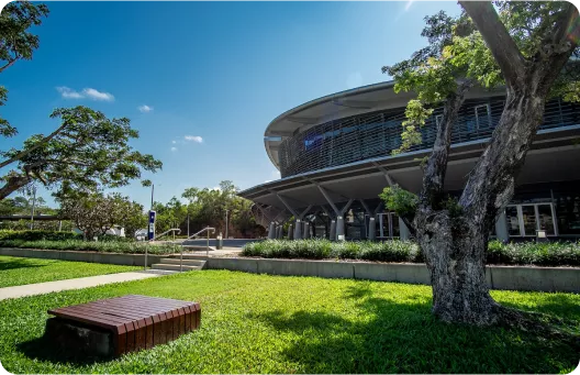 A modern building with a curved roof, surrounded by greenery and trees, features a wooden bench in the foreground