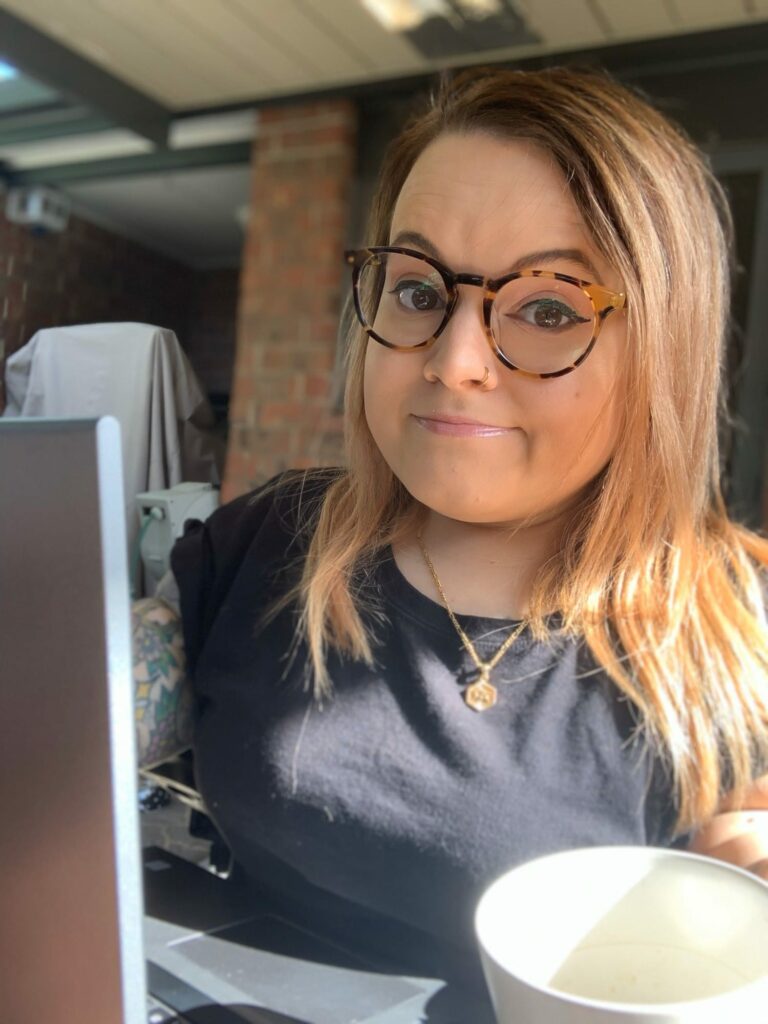 A young woman with long hair and glasses sitting at her laptop with a hot drink. She is wearing a black t-shirt with a gold necklace.