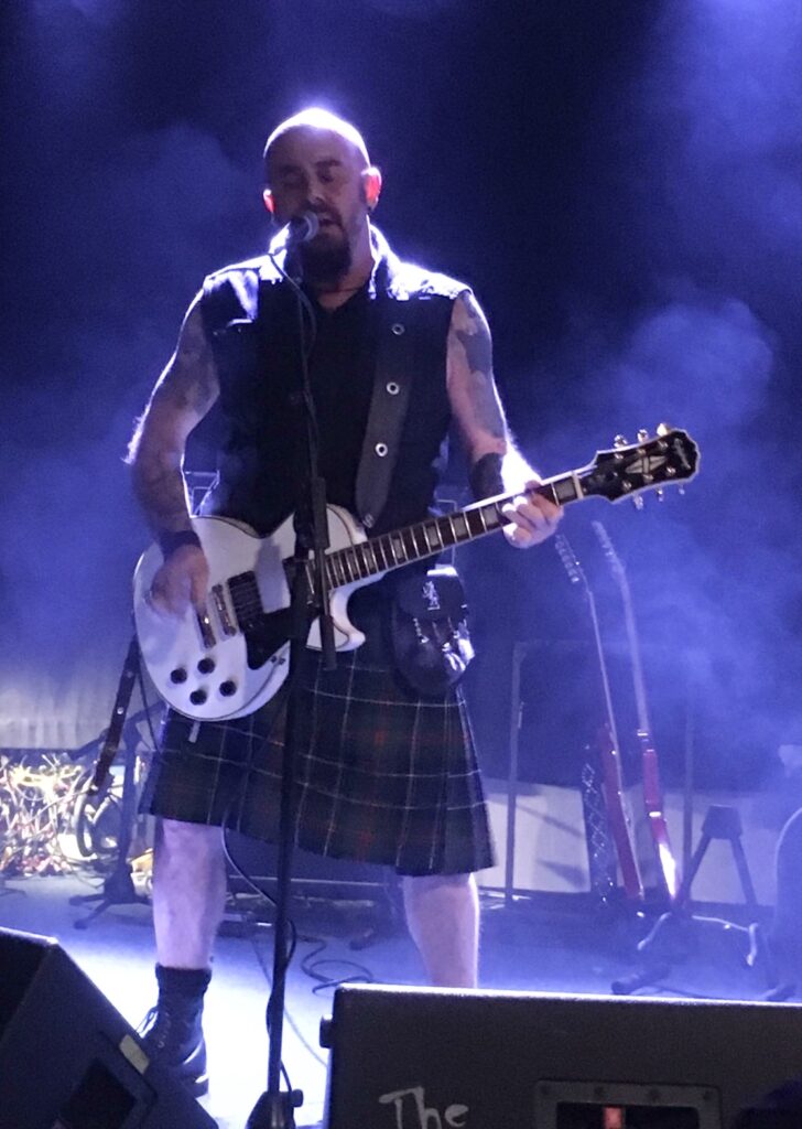 A man singing and playing electric guitar on stage. He has tattoos on his arms and wears a kilt.