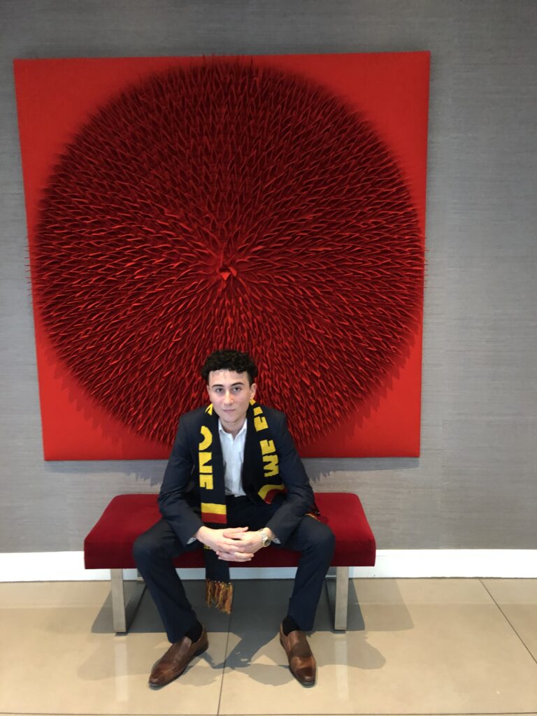 A man in a suit and a football scarf sits in front of a red artwork.