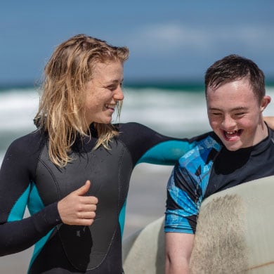 A young man and a young woman standing on the beach. They wear wetsuits and the man is carrying a surfboard.