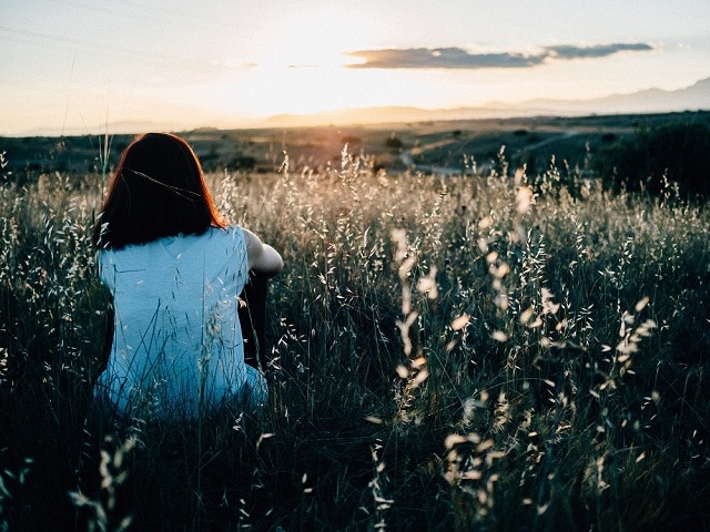 A girl sitting in a field looking out to the sunset.