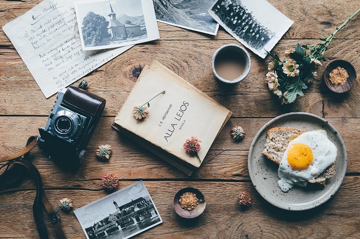 A wooden table with a camera, black and white photos, a handwritten note, a book, an egg on toast, a coffee and some flowers