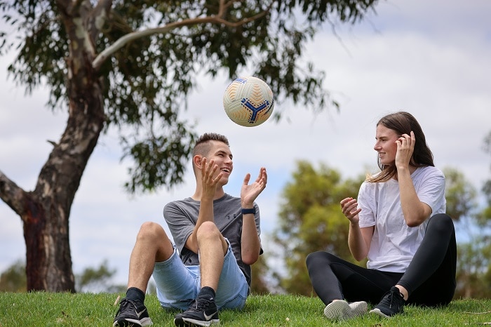 A man and a woman sitting in a park smiling, the man is throwing a soccer ball in the air