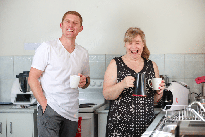 A man and a woman standing in a kitchen holding cups of tea. They are looking at the camera and laughing.