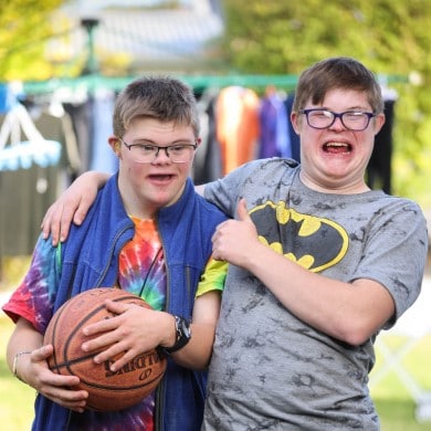 Two boys in a backyard with their arms over each others' shoulders. One is holding a basketball.
