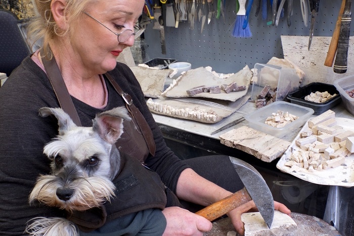 Louise Marson in her studio holding a hammer to a slab of rock with her assistant dog Penny in her lap