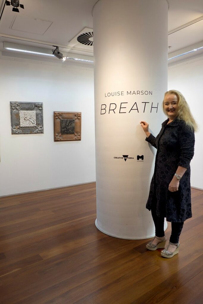 Louise Marson standing in front of a pillar that's branded with her exhibition name 'Breath' at the Dax Centre with two of her works displayed in the background. 