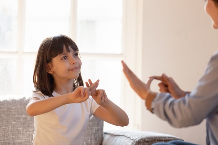 A young girl with brown hair smiles whilst communicating with an adult using sign language.