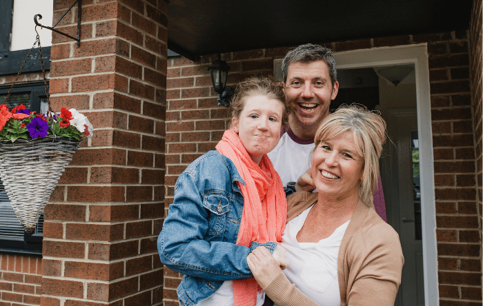 A smiling mother and father stand on the front porch of their home with their daughter who has a disability.