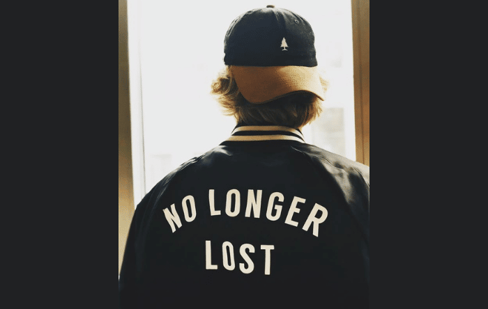 A young man wearing a backwards cap and jacket with the words 'no longer lost' stares out of a window.