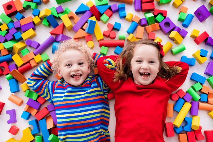 Two children lying on the ground in amidst colourful building blocks, smiling for a photo.